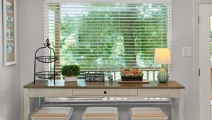 Una ventana con persianas horizontales, una pequeña planta en maceta, una bandeja con aperitivos, un letrero decorativo y una lámpara sobre una mesa de madera.