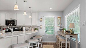Una cocina moderna con armarios blancos, un microondas de acero inoxidable y un horno empotrado. Hay tres lámparas colgantes, una ventana con persianas y una pequeña mesa con una lámpara.