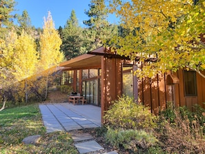 Une cabane moderne en bois, dotée d’une porte en verre et d’un patio avec une table et des chaises, le tout entouré du feuillage automnal.