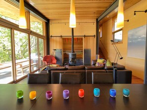 Un intérieur moderne avec un plafond en bois, de grandes fenêtres, un ensemble de canapés, une cheminée et une table agrémentée de verres colorés.
