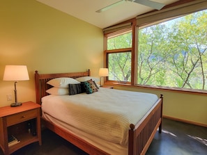 Een slaapkamer met een groot raam, een bed met veel kussens, een nachtkastje met een lamp en een klein dressoir.