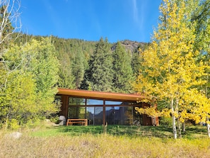 Une cabane moderne dotée de grandes fenêtres, d’une terrasse en bois et offrant une vue sur une chaîne de montagnes boisée.
