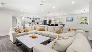 A modern living room with a sectional sofa, a coffee table, and a kitchen in the background. - Grand Retreat: Luxury Vacation for Large Groups (Fayetteville)