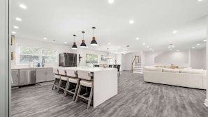 A modern kitchen with a central island, bar stools, pendant lighting, and a white sofa in a spacious living area. - Grand Retreat: Luxury Vacation for Large Groups (Fayetteville)