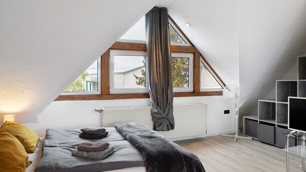 A bedroom with a sloped ceiling, a bed with pillows, a window with curtains, and a radiator. - Studio Apartment 'Vogelsberger Bett Die 2' with Wi-Fi (Alsfeld)