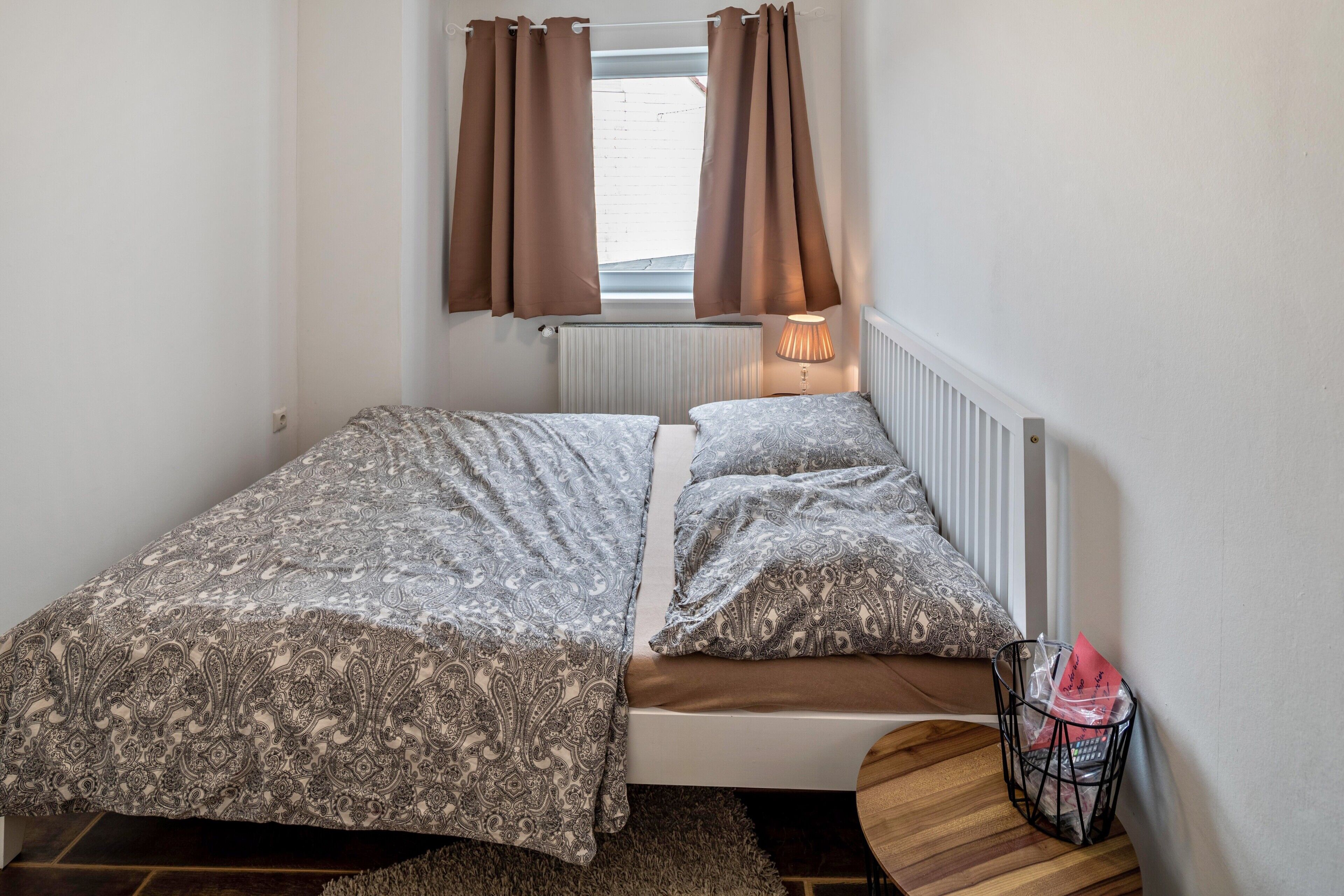 A bedroom with a bed, a radiator, curtains, a lamp, and a small round table with a basket.