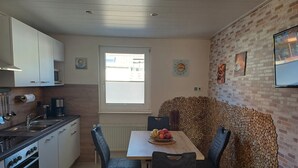 Een keuken met witte kasten, een fornuis en een raam. Er is een eettafel met stoelen en een schaal met fruit.