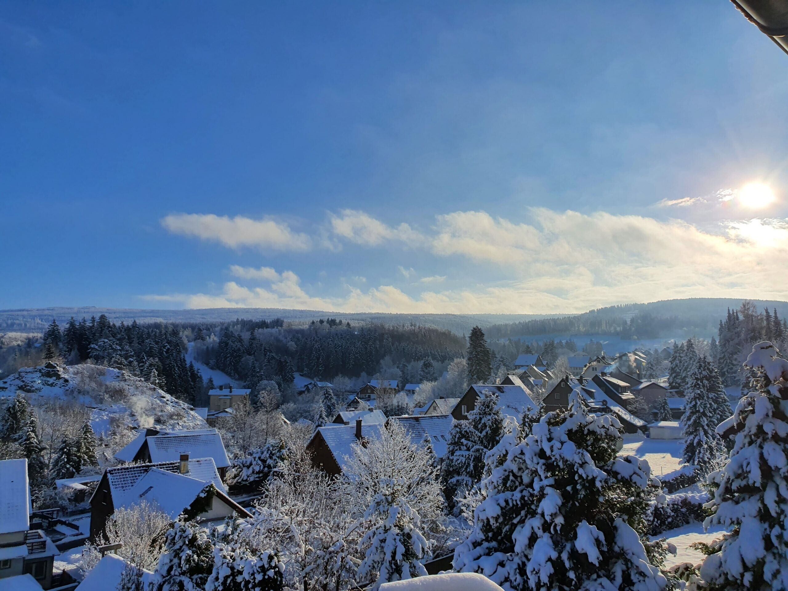 Un pueblo cubierto de nieve con casas y árboles, cielo despejado y una vista distante de colinas.