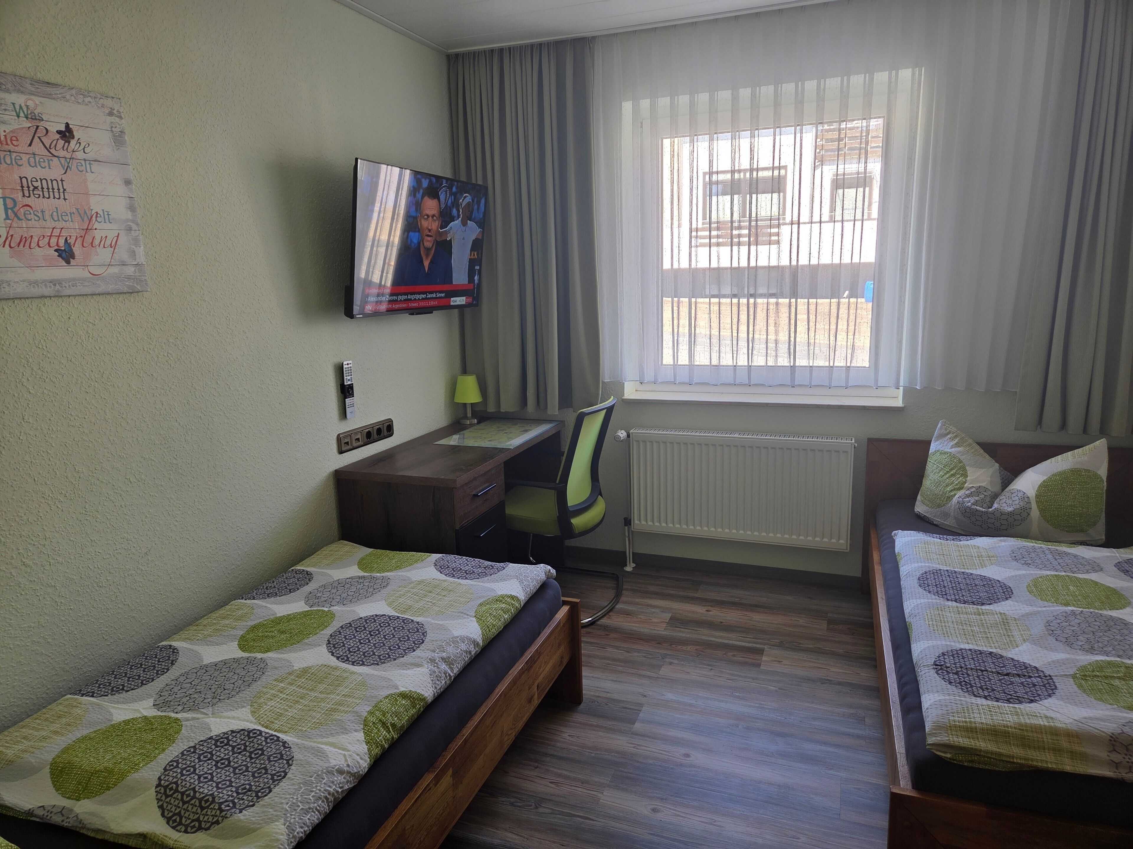A hotel room with two single beds, a desk, a chair, a TV, and a window with curtains.