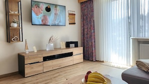 Un salon avec un meuble TV en bois, un téléviseur à écran plat, une radio et un panier de fruits posé sur une table ronde.