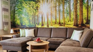Een woonkamer met een hoekbank, een klein houten tafeltje met fruit, en een wanddecoratie van een bos.