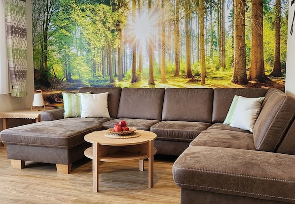 Un salon avec un canapé d’angle, une petite table en bois sur laquelle sont disposés des fruits, et une fresque représentant une forêt sur le mur.