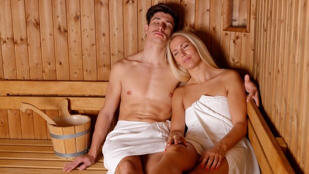 Een houten sauna met banken, een houten emmer en een persoon met een handdoek.