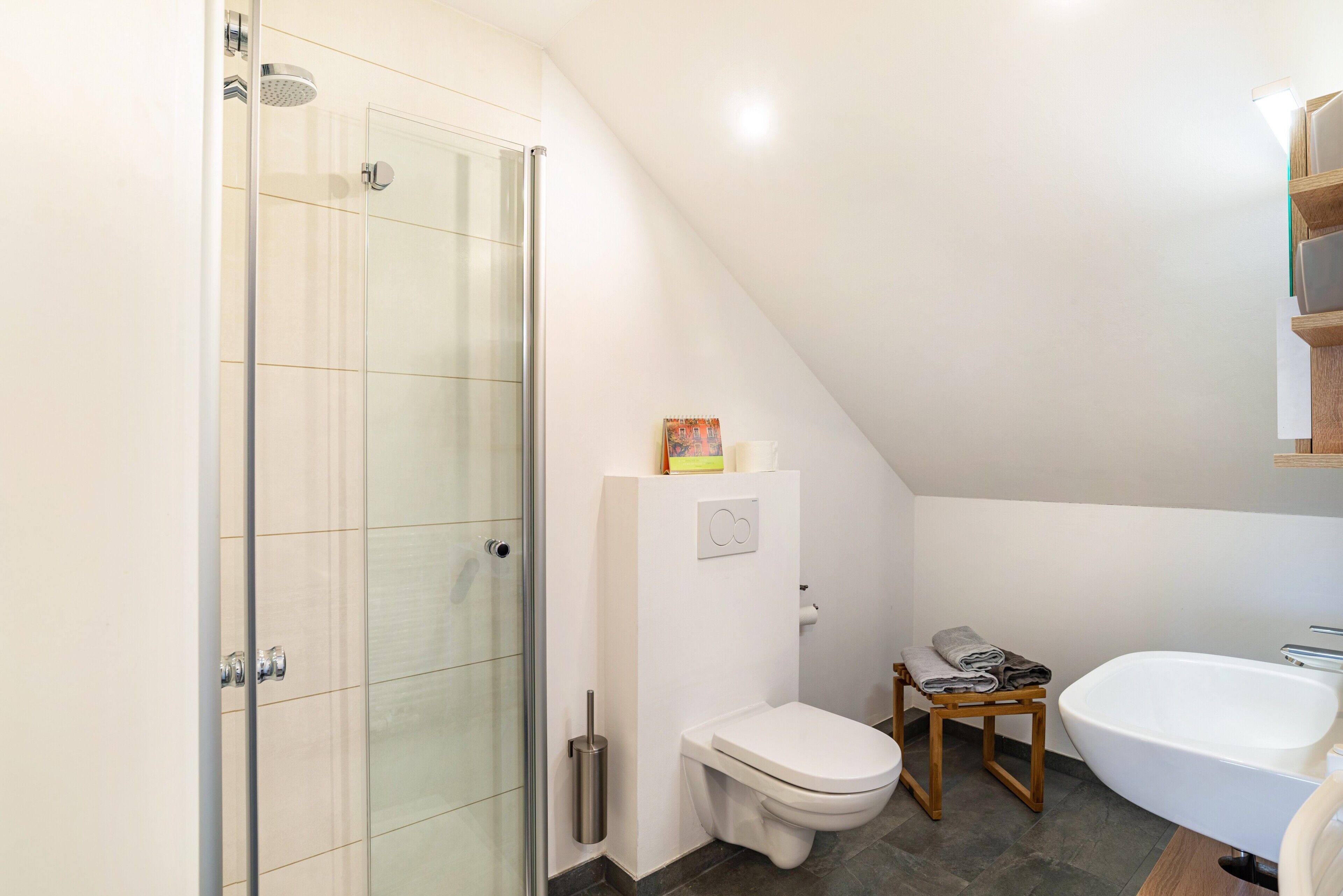 Ein Badezimmer mit Duschkabine aus Glas, einer weißen Toilette, einem hölzernen Hocker und einer weißen Badewanne.
