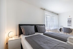 Een hotelkamer met twee bedden, een nachtkastje met lamp, een stoel, een raam met gordijnen, en een muur met een religieuze poster.