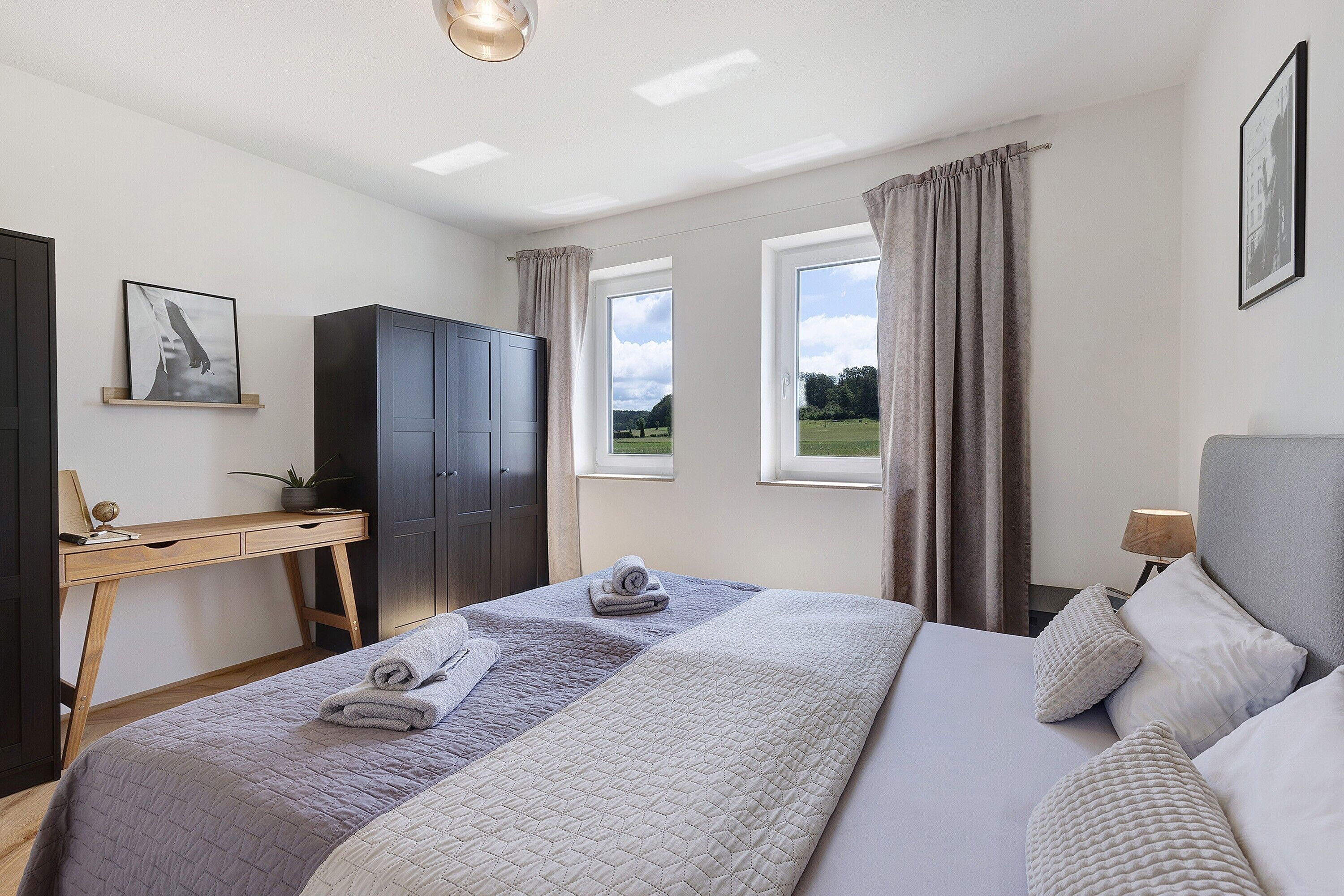 Ein Schlafzimmer mit Bett, Schreibtisch, Kleiderschrank und zwei Fenstern mit Vorhängen.