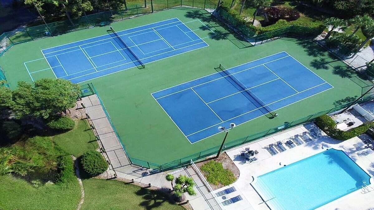 Vista aérea de un complejo de canchas de tenis con dos canchas, una piscina y zonas verdes circundantes.
