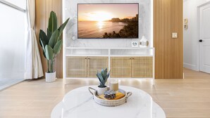 A modern living room with a large flat-screen TV mounted on a white marble wall, a wooden accent wall, a large plant, and a round coffee table with decorative items. - High Floor 2BR/2BA+Den 4beds Partial Ocean View + 1 Parking 2 min from the Beach (Honolulu)