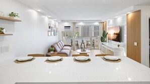 A modern living room with a dining area, white walls, and a large TV. - High Floor 2BR/2BA+Den 4beds Partial Ocean View + 1 Parking 2 min from the Beach (Honolulu)