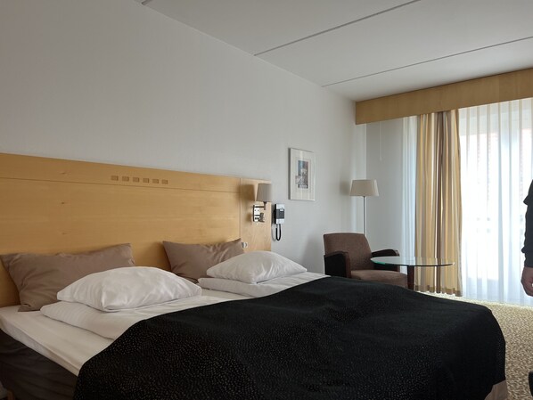 Habitación de hotel con cabecera de madera, cama con ropa de cama blanca y beige, manta de color negro, una mesita con lámpara y una silla.