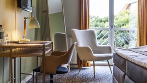 Habitación de hotel con cama, escritorio con lámpara, dos sillas, un espejo grande y ventana con cortinas.