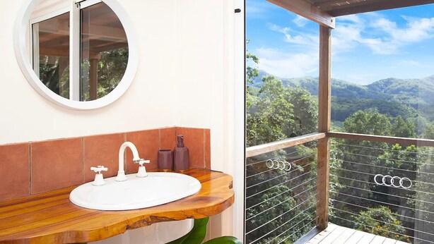 Ein Badezimmer mit rundem Spiegel, weißem Waschbecken und Holzschrank. Es besitzt einen Balkon mit Blick auf grüne Hügel und einen klaren Himmel.