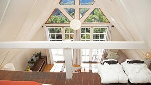 Ein gemütliches Schlafzimmer unter dem Dach mit Holzboden, einem Bett mit weißer Bettwäsche, einem kleinen Tisch mit Lampe und Blick ins Grüne durch große Fenster.