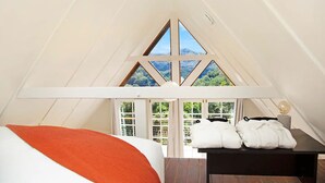 Ein Schlafzimmer mit schrägverdachter Decke, einem Bett mit orangefarbener Tagesdecke, einem Nachttisch mit Lampe und Blick ins Grüne durch die Fenster.