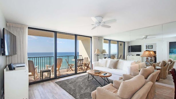 A spacious living room with a large sliding glass door leading to a balcony with a view of the ocean. There is a ceiling fan, a sofa set, and a coffee table.
