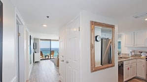A hallway leading to a balcony with a view, white walls, and a kitchen with white cabinets and a stainless steel refrigerator.