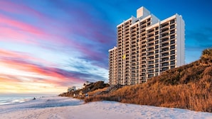 Un hôtel de grande hauteur doté de nombreux balcons, situé à proximité d’une plage, avec un ciel dégagé et un coucher de soleil.
