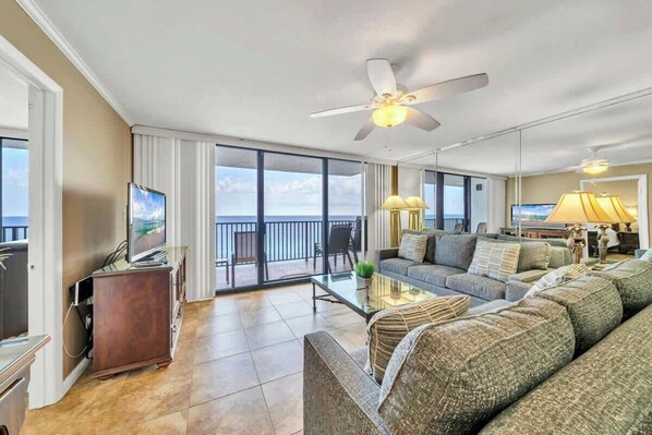 A spacious living room with a large sofa set, a coffee table, and a television. There is a balcony with a view of the ocean.