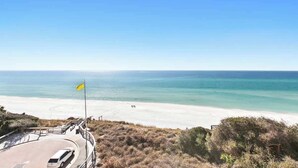 A coastal road leading to a beach with a flag, clear skies, and calm sea.