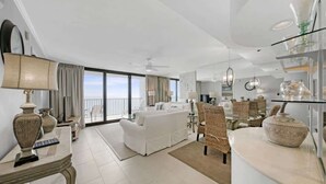 A spacious living area with a white sofa, dining table, and chairs, a balcony with a view, and a modern kitchen area.