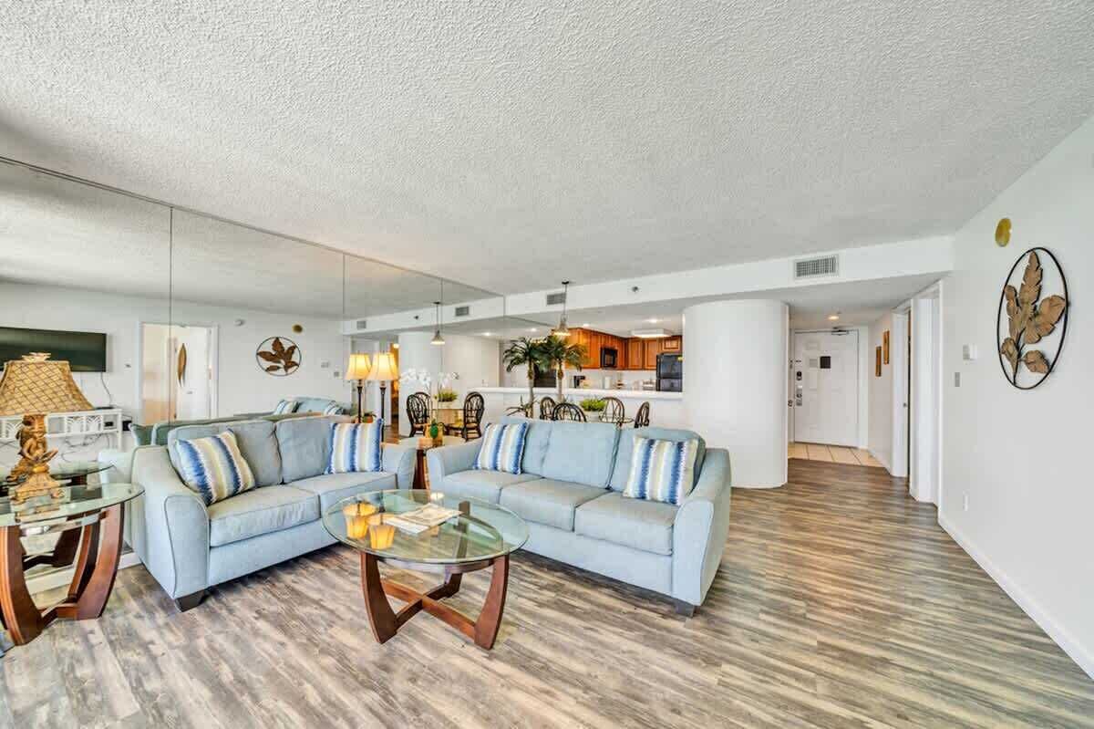 A spacious living area with a light blue sofa, a glass coffee table, and a kitchen in the background.