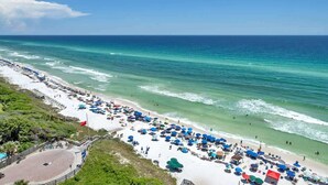 A beach with rows of umbrellas and sun loungers, a clear view of the ocean, and a flag on a flagpole. - One Seagrove Place 1306 (Santa Rosa Beach)
