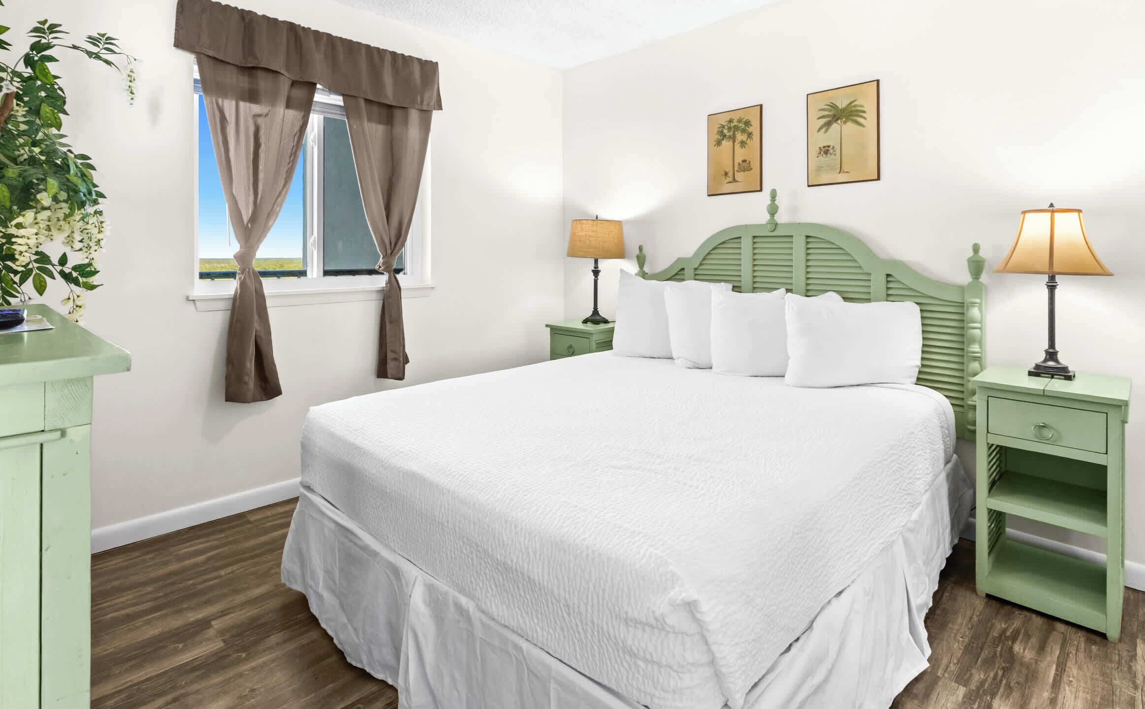A bedroom with a large bed, two bedside tables with lamps, a window with curtains, and two wall-mounted pictures.