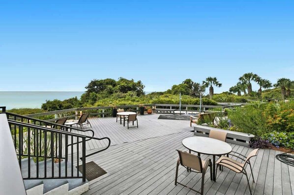 A wooden deck with outdoor seating, overlooking a body of water and lush greenery. - One Seagrove Place 1706 (Santa Rosa Beach)