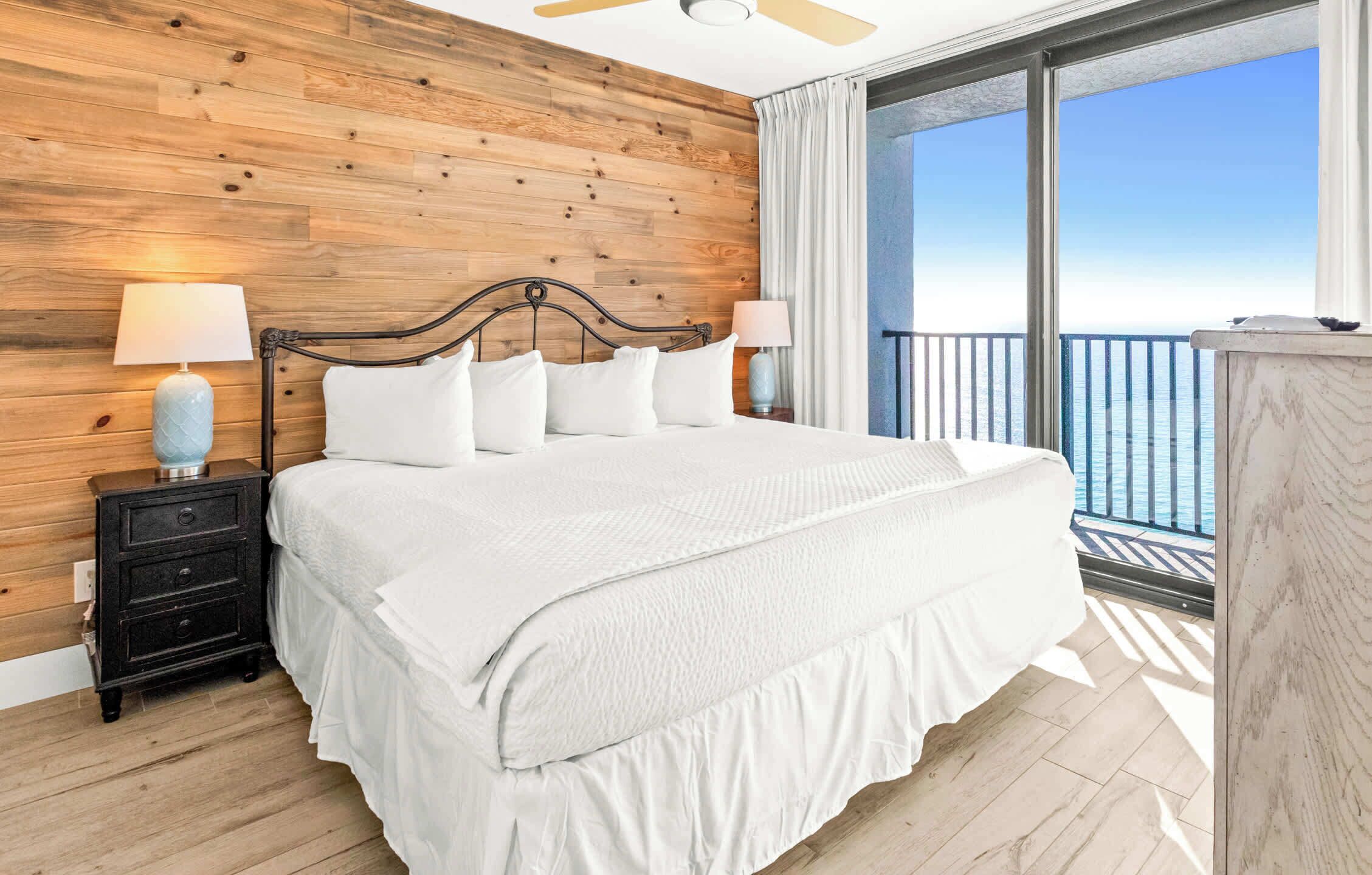 Une chambre à coucher avec une tête de lit en bois, un grand lit avec une literie blanche, deux tables de chevet avec des lampes et une vue sur la mer depuis un balcon.