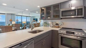 A modern kitchen with stainless steel appliances, a large window, and a dining area with wicker furniture. - One Seagrove Place 1706 (Santa Rosa Beach)