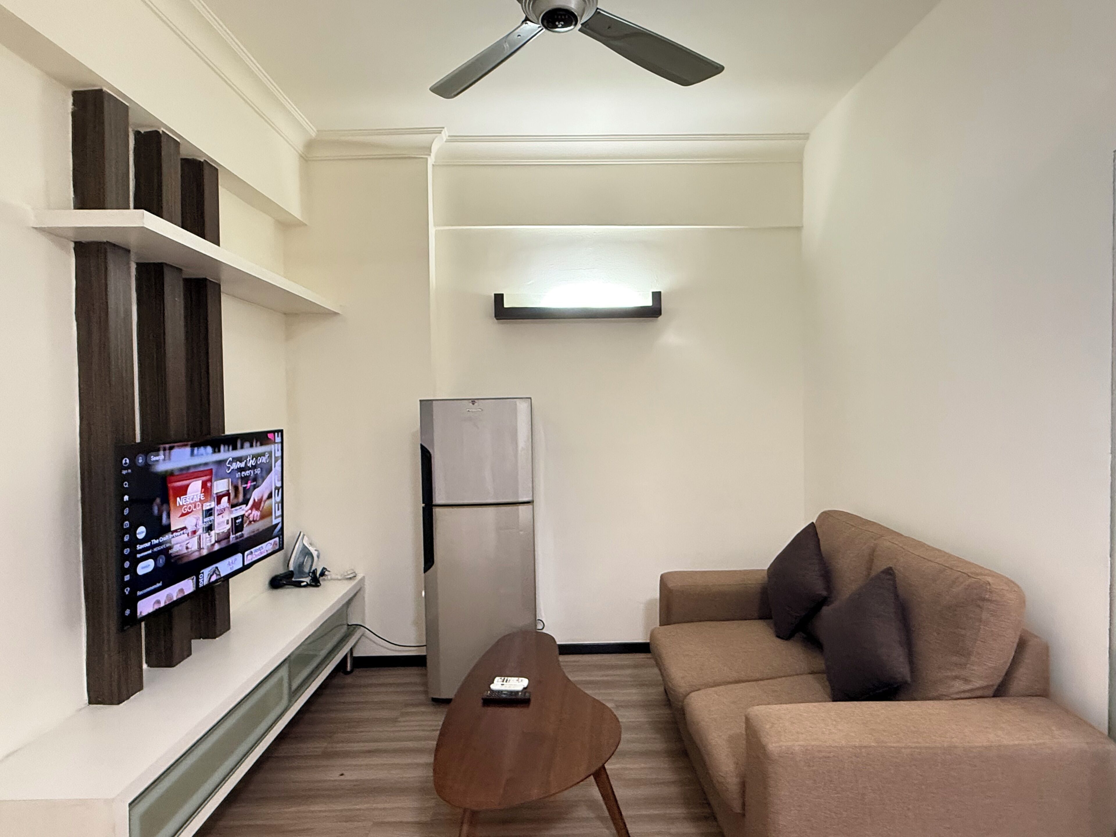 A living room with a flat-screen TV mounted on the wall, a refrigerator, a sofa, and a wooden coffee table.