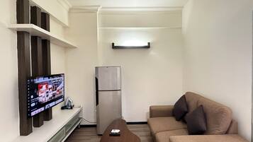 A living room with a flat-screen TV mounted on the wall, a refrigerator, a sofa, and a wooden coffee table.