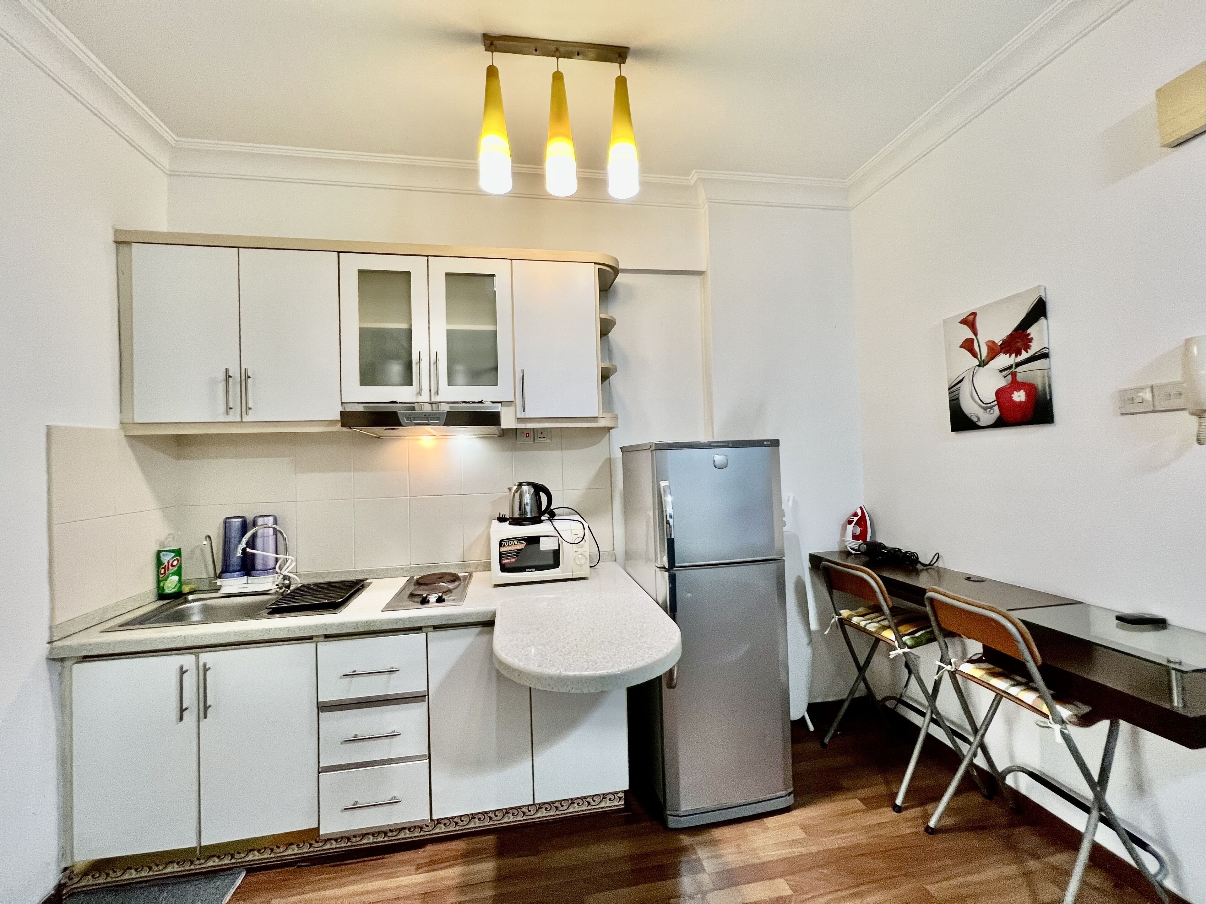A compact kitchen with white cabinets, a microwave, a refrigerator, and a small dining area with a chair.