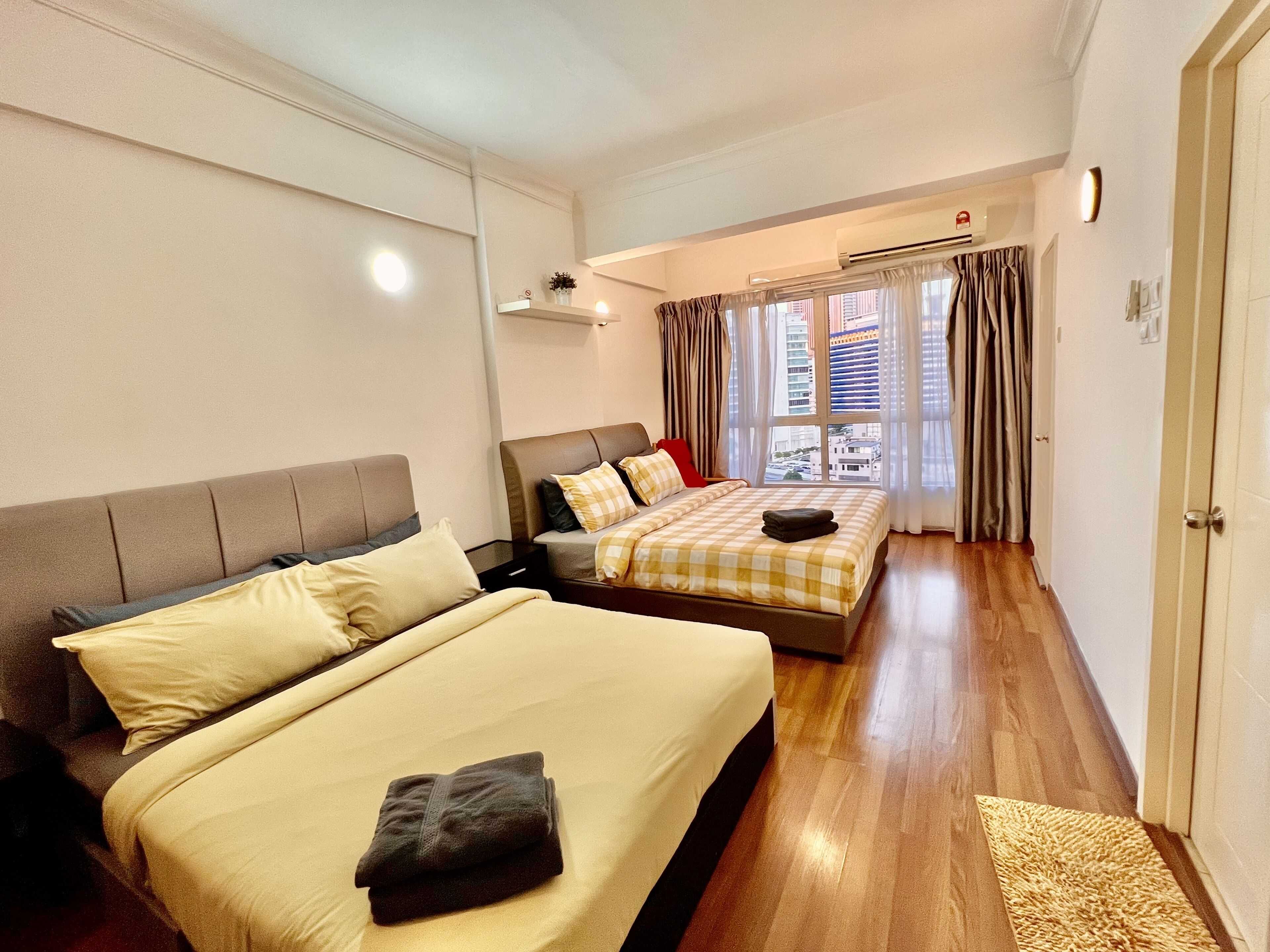 A hotel room with two beds, a window with curtains, a door, and a small table with a lamp.