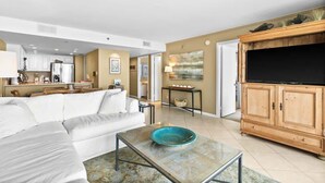 A living room with a white sofa, a glass coffee table with a blue bowl, a wooden TV stand, and a kitchen in the background. - One Seagrove Place 0405 (Santa Rosa Beach)