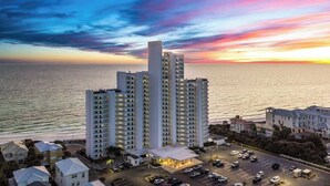 A high-rise hotel with multiple floors, a parking lot, and a view of the ocean at sunset. - One Seagrove Place 0405 (Santa Rosa Beach)