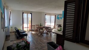 Un vaste espace de vie comprenant un canapé, une table à manger et des chaises. On aperçoit un piano et une poubelle au premier plan.