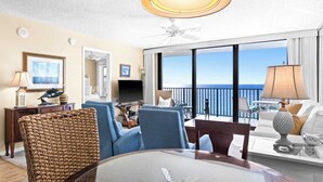 A spacious living room with a large window offering a view of the ocean, a ceiling fan, and a dining area with a table and chairs.