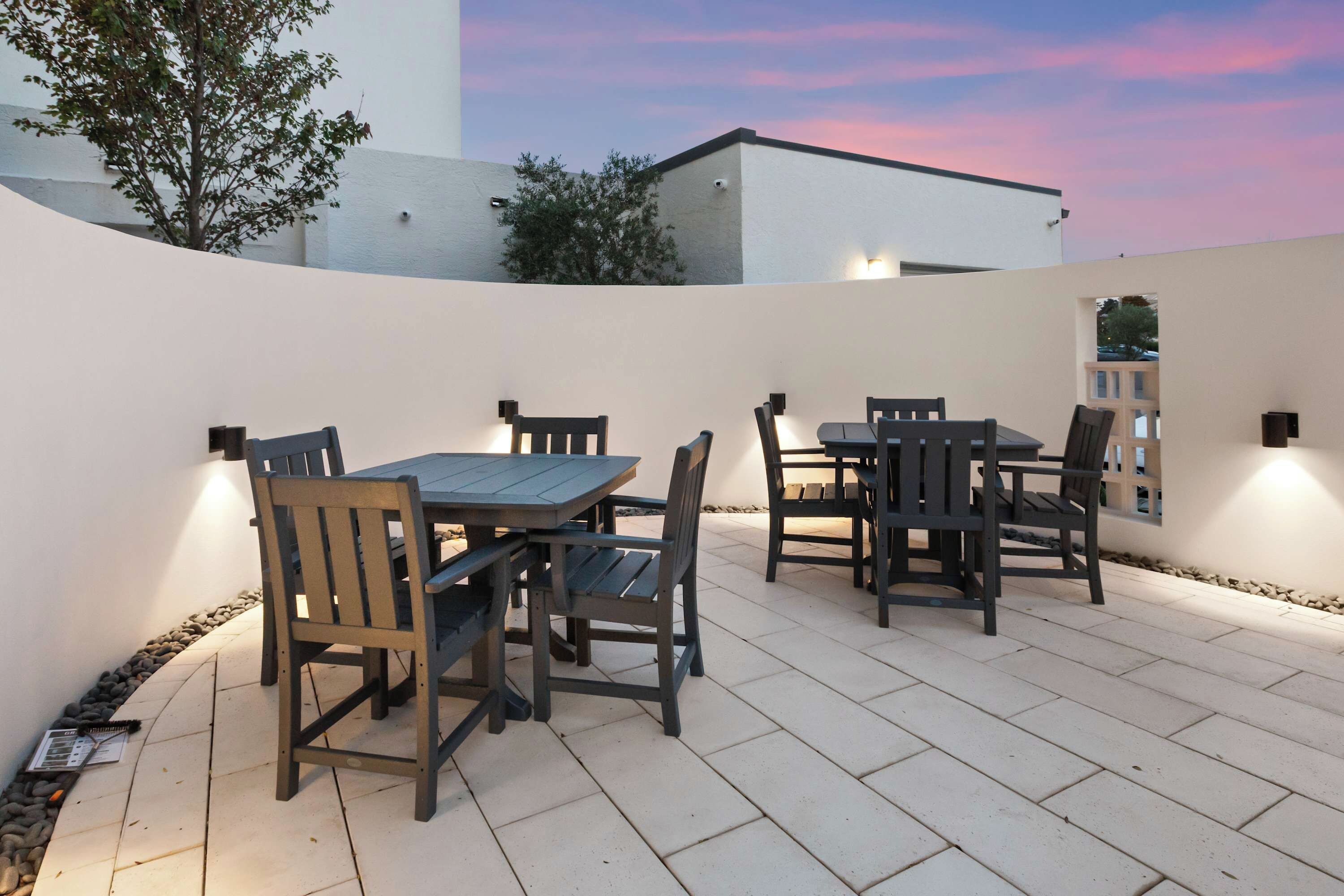 An outdoor patio with a table and chairs, surrounded by a white wall and a view of a building and trees.