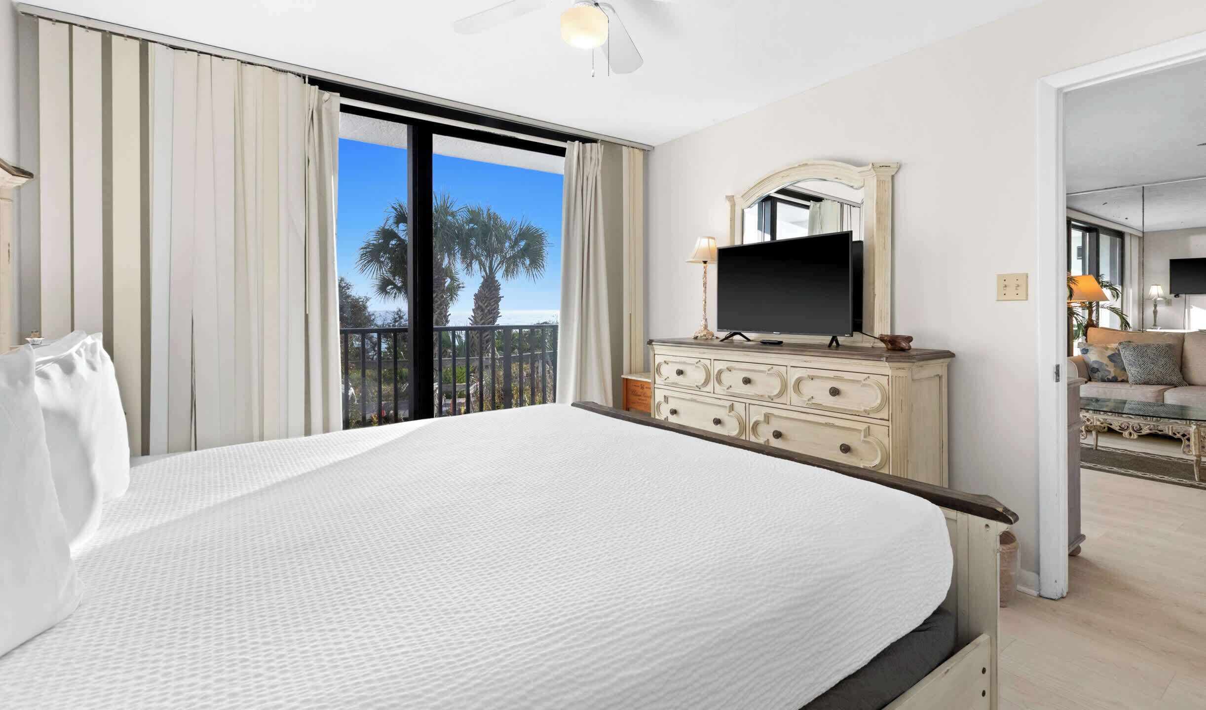 A hotel room with a large bed, a dresser with a mirror, a flat-screen TV, and a balcony with a view of palm trees and the ocean.